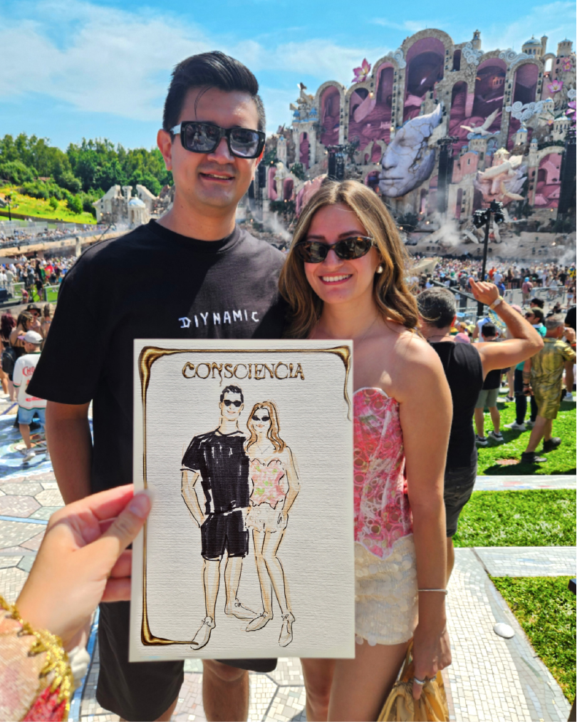 My hand holding a live illustration I made of the couple standing behind it, in front of the Mainstage at Tomorrowland